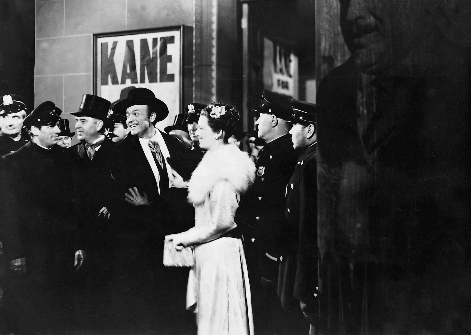 Visuel Citizen Kane : un homme souriant en manteau et chapeau, accompagné d’une femme en robe élégante, avance parmi des policiers et des journalistes.